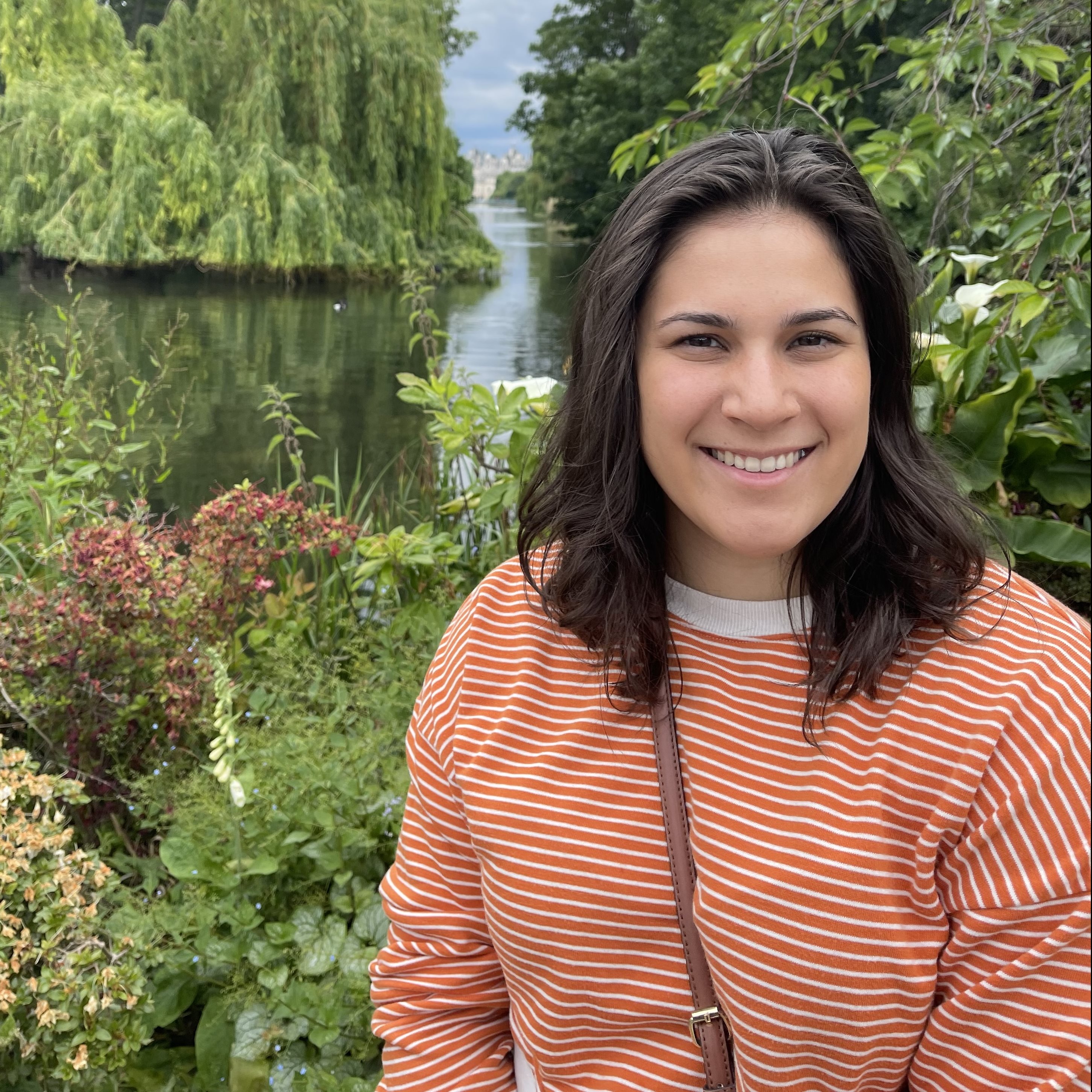 Woman in orange and whit horizontally striped shirt poses smiling in front of a beautiful lake with trees and bushes around it.