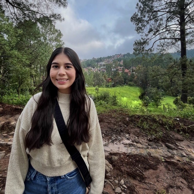 Woman with long brown hair in a cream sweater smiling and posing in front of a beautiful green hill with trees and a small town in the background.