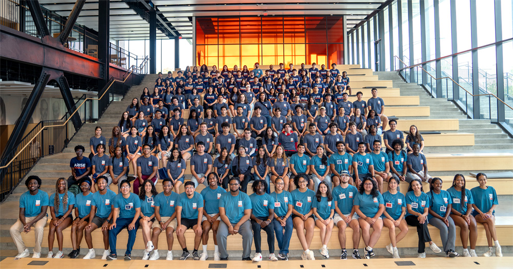 University students in matching blue t-shirts sit smiling at the camera on tiered bleachers.